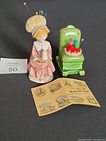 Front angle view of vintage porcelain pin cushions and needle pack. Girl figurine pin cushion has pins on bonnet. Green chair pin cushion has red padded cushion with pins.