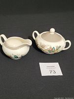 Photo showing the creamer and sugar bowl with lid side by side on a black surface with auction lot number card.