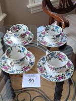 Four vintage porcelain tea cups and matching saucers with colorful floral patterns and gold rims, displayed on glass with wooden chair background.