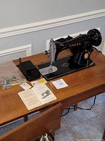 Front angled view of Singer sewing machine on cabinet with pedal and sewing accessories including needles and instruction manual.