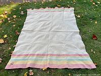 Full view of the white wool blanket spread on grass showing the pastel stripe edges and scalloped edge design.