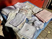Wide view of multiple folded antique estate linens with various textures, embroideries, and lace on a table outdoors.