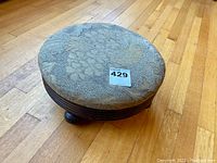 Top view of round foot stool showing floral upholstery and three turned legs on hardwood floor.
