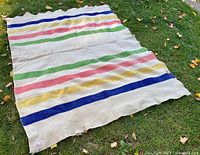 Full view of the cream wool blanket with blue, yellow, pink, and green horizontal stripes spread out on grass, showing overall pattern and condition.