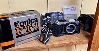 Photo of Konica FT-1 Motor camera body with case, original box, colorful strap, and instruction manual all displayed on a wooden surface.