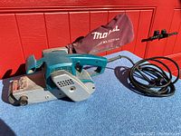 Side view of Makita belt sander with attached power cord and dust collection bag on blue table.