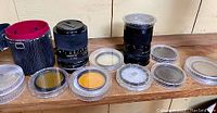 Various vintage camera lenses, filters, and carrying case arranged on a wooden surface.