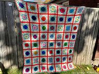 Full view of the vintage hand knit afghan blanket hanging outdoors, showing all colorful squares with red border grid.