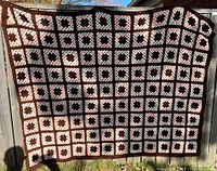 Full view of the knitted afghan blanket showing the square patch pattern in brown, pink, and white, hung over a fence outdoors.
