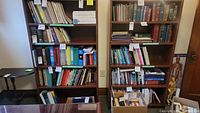 Two medium brown wooden bookshelves filled with books. Shelves have 5 levels each, with various books placed on them. The bookshelves are standing side by side against a wall.