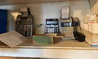 Wide view of all calculators and numbering machine on shelf, showing various models and paper rolls.