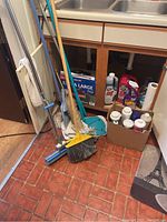 Photo showing mop, broom, dustpan, box of extra large trash bags, and assorted cleaning chemical bottles under kitchen sink.