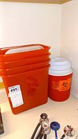 Photo showing three large orange sharps disposal containers stacked with white lids and a small orange cylindrical sharps container with biohazard label beside them.