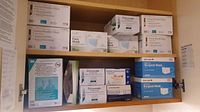 Overall view of multiple boxes of masks including Earloop Procedure Masks, Surgical Masks, and N95 respirators along with gloves and face shield stored inside a cabinet.