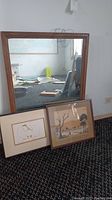 Full view of mirror leaning against a wall with the two framed artworks placed in front of it on carpeted floor.
