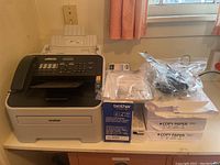 Brother Intellifax 2840 fax machine with handset, control panel, and paper tray, placed on a table with toner cartridge box and multiple copy paper packs beside it.