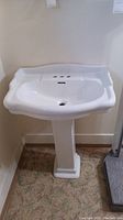 Front view of white ceramic pedestal bathroom sink showing basin, three faucet holes, and pedestal base on carpeted floor.