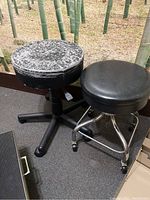 Two black swivel stools with round seats in front of a window showing an outdoor bamboo scene. One stool seat is heavily worn and damaged; the other stool seat is in better condition. Bases differ, one black plastic with foot lever, one chrome metal with caster wheels.
