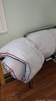 White quilted bedspread with red and blue trim folded on metal frame, queen size.