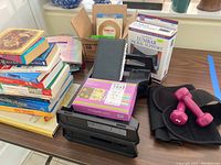 Image showing stacked books, lumbar sacral support in box, pink dumbbells, black lumbar support belt, pack of blank cards, and black desktop organizer arranged on a table.