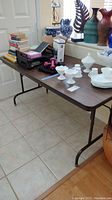 Full view of folding table with various unrelated items placed on top including books, boxes, and white glassware, shown to illustrate table condition and size.