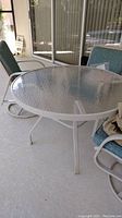 Full view of round glass topped patio table with white metal frame and outdoor chairs partially visible.
