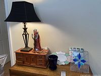 Lamp, wooden jewelry box with drawers, hand-shaped jewelry holder with costume jewelry, dark blue vase, ceramic plates, and leaded glass panel displayed on top of a wicker chest against a white wall.
