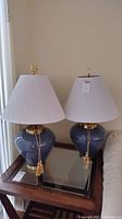 Pair of matching table lamps with blue ceramic urn-shaped bodies, brass-colored round bases, white pleated fabric shades, and beige hanging tassels, shown side by side on a wooden glass-top table near a beige wall and window.