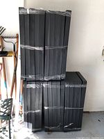 Five black plastic stackable garage shelving units, sealed and brand new, stacked vertically and secured with clear plastic wrap.