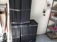 Five black plastic shelving units stacked and shrink-wrapped in clear plastic, showing front and side view of units pressed against a wall next to garden tools.