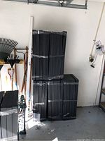 Sealed bundles of black plastic shelving units stacked in garage with garden tools visible.