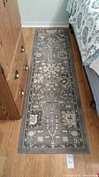 Rectangular runner rug with gray floral geometric pattern next to wooden dresser and bed, approx. 7 ft long
