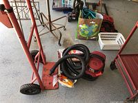 Overview showing shop vac with coiled black hose on top and spare vacuum bags near metal convertible hand truck, castors and wheels visible.