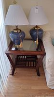Wood side table shown beside sofa with two blue lamps with brass accents on top. The wood finish is dark brown with a beveled glass insert on the top surface.
