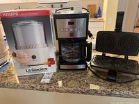 Three kitchen appliances on a countertop: a Krups ice cream maker in box, a Kenmore drip coffee maker with glass carafe, and a Chefs Choice PizzellePro waffle maker opened to show the patterned cooking plates.