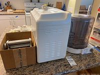 Three small kitchen appliances on a kitchen counter: Welbilt bread machine, pasta maker in box, Oster Carousel Rotisserie