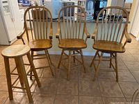 Three wooden swivel counter stools with armrests and spindle backrests plus one additional stool with round seat without armrests, all placed on tiled floor in kitchen setting.