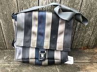 Front view of laptop bag showing vertical striped seatbelt material in neutral tones with flap and seatbelt latch clasp.