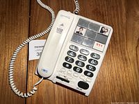 Photo of the Serene corded desk phone displaying the handset, numeric keypad with oversized black buttons, and six memory dial buttons with photo slots, including a 911 button. Phone and cord are visible on a dark wooden surface.