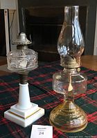 Two vintage oil lamps placed on a table with a plaid cloth; one white, one gold-toned, one with the hurricane glass chimney and one without.