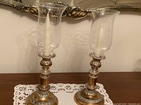Two antique silver-toned candle holders with glass shades on lace doily, one with a taper candle inserted
