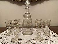 Photo of central decanter surrounded by six matching glasses, all featuring white and gold enamel floral decorations on clear blown glass.