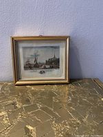 Frontal photo of the framed vintage lithographic print showing harbor scene with boats and buildings under a partly cloudy sky. The gilded wood frame is visible with decorative beading on the inner edge.