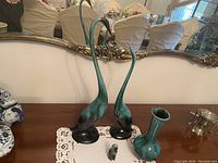 Photo showing two tall crane bird figurines, one vase, and a small bird figurine on a wooden surface with a large framed mirror in the background.