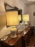 Pair of off-white ceramic table lamps with 3D cherry blossom detailing on bases and beige lamp shades, placed on a polished wooden ornate furniture below a decorative mirror.