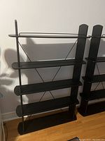 Two black mid-century modern shelves, metal and wood, showing front view with cross-bracing and rounded edges on wooden shelves.