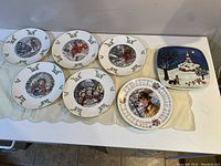Seven round and one square vintage Royal Doulton collectible plates with detailed winter and holiday scenes, gold trim, and holly borders displayed on table.