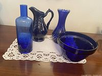 Photo of five cobalt blue glass items: a tall bottle, an ornamental pitcher, a flared vase, and a small bowl all placed on a lace doily on a wooden surface.