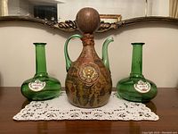 Front view of three vintage collectable bottles on a doily, showing the elaborate map design and lion emblem on the large decanter with two green bottles labeled 'Sherry' and 'Port'.