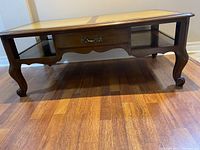 Front view of vintage mahogany coffee table showing the leather top, central drawer with metal handles, and curved legs, placed on wooden flooring.